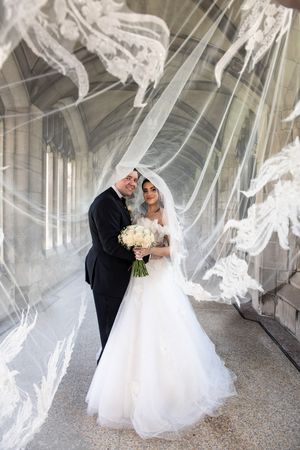 Wedding couple embracing under billowing lace veil in ornate stone corridor holding white bouquet.