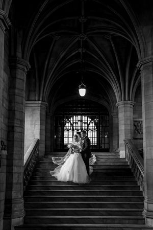Bride and groom silhouetted on grand staircase under Gothic arched ceiling in black and white.