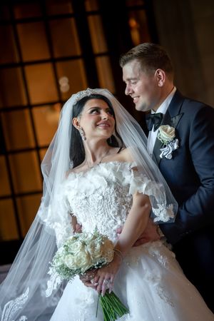Smiling bride in off-shoulder lace gown with tiara and groom in tuxedo sharing tender moment.