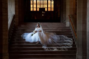 Elegant bride in cathedral-length lace veil seated on grand stone staircase bathed in golden light.
