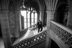 Bride and groom walking hand in hand on ornate balcony staircase in black and white photo.