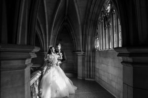 Bride and groom in intimate embrace beside Gothic arched window in dramatic black and white portrait.