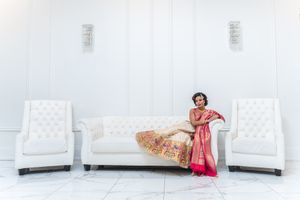 Indian bride in vibrant pink and gold saree poses gracefully on white tufted sofa in bright venue.