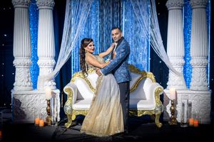 Indian couple poses elegantly before blue sequined backdrop with white ornate thrones at wedding reception.