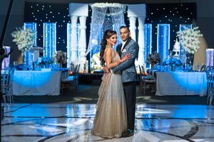 Indian bride and groom embrace on dance floor before stunning blue-lit floral wedding reception decor.