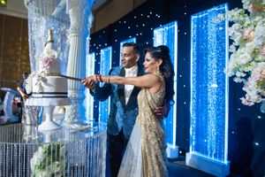 Indian bride and groom cut tiered white wedding cake before glowing blue illuminated reception backdrop.