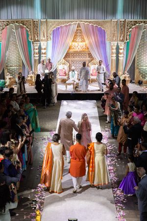 Indian bride walks down petal-lined aisle toward ornate mandap during traditional Hindu wedding ceremony.