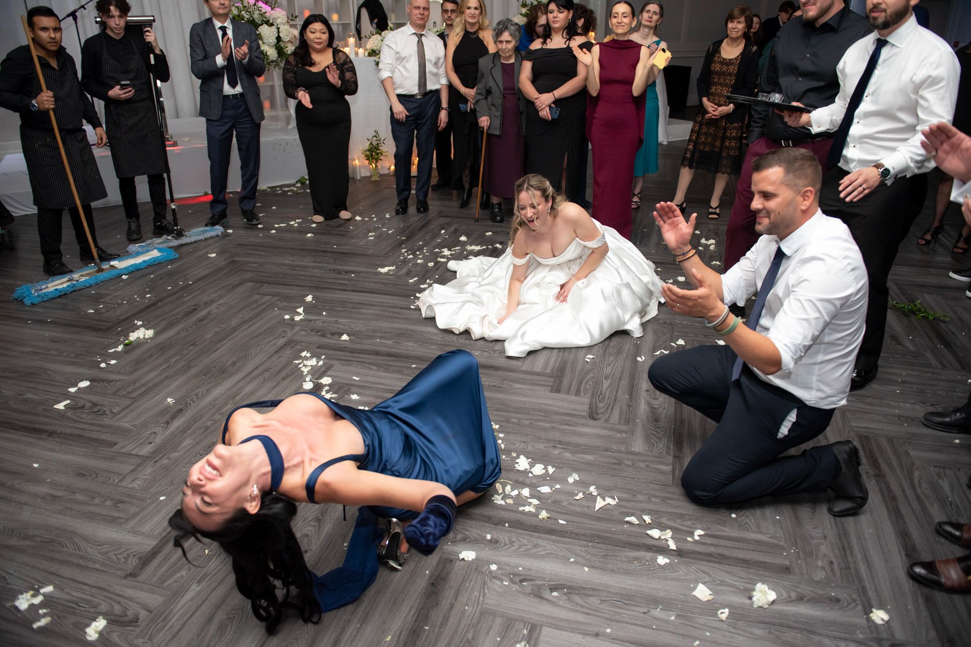 Lively wedding reception limbo dance with guests cheering as man bends backward on dance floor.