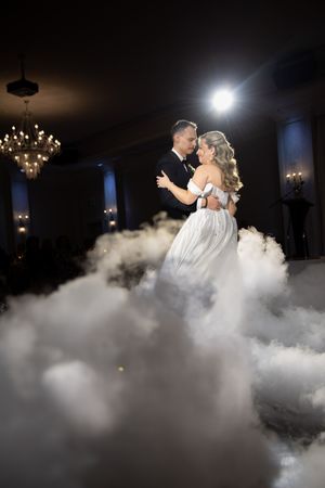 Bride and groom share romantic first dance surrounded by dramatic fog effect in dimly lit ballroom.
