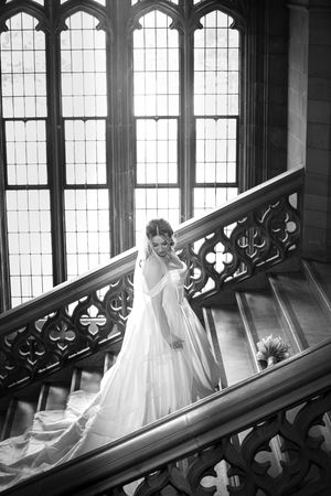 Elegant black and white portrait of bride in wedding gown on ornate staircase by tall windows.
