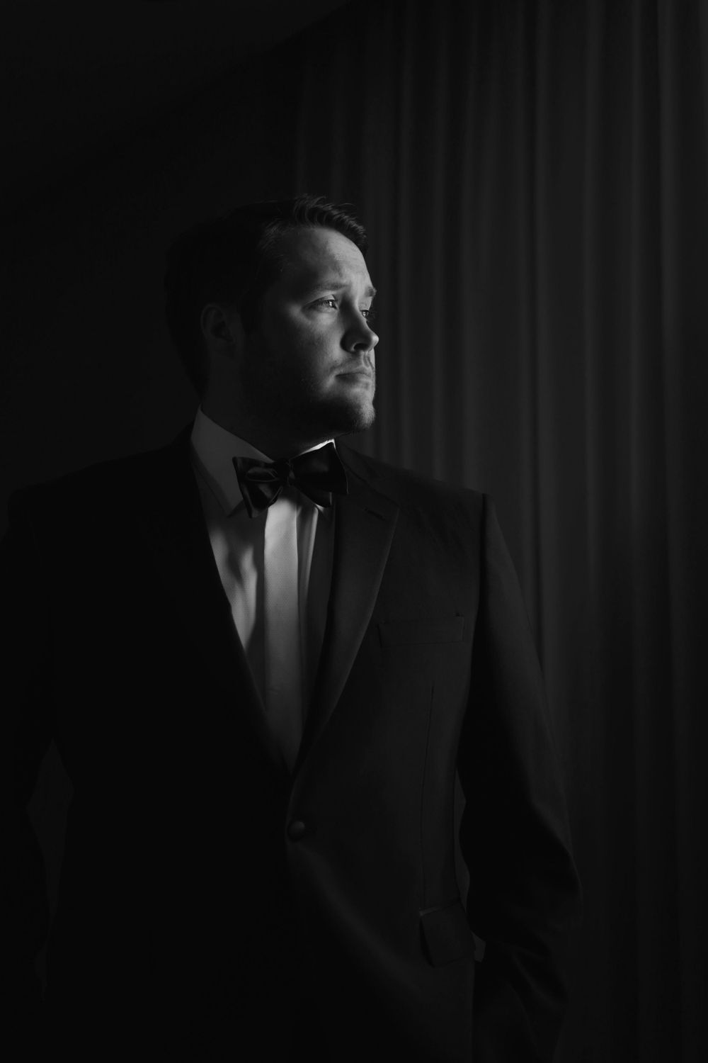Black and white dramatic portrait of a man in a tuxedo and bowtie gazing to the side in moody lighting.