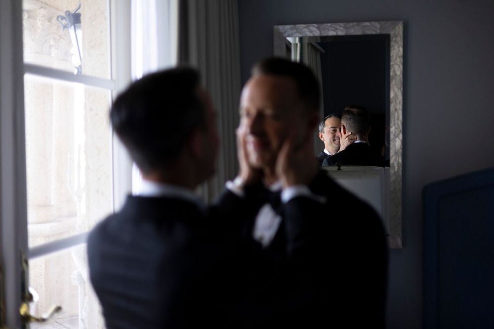 Two people in black suits stand near a window in a dimly lit room with a mirror on the wall.