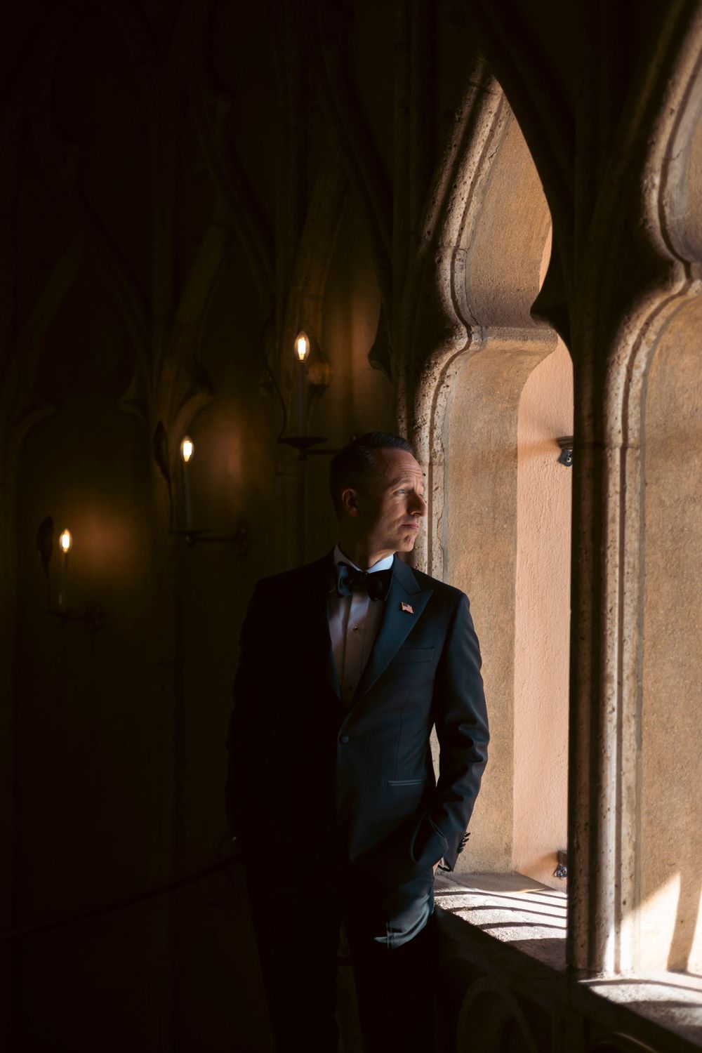 A groom in a dark suit stands thoughtfully in an archway with dramatic lighting and stone architecture.
