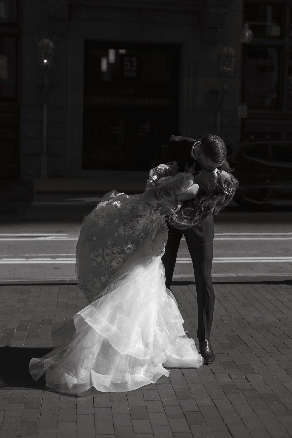 A black and white photograph of a couple in wedding attire sharing a romantic dip dance pose on a city street.