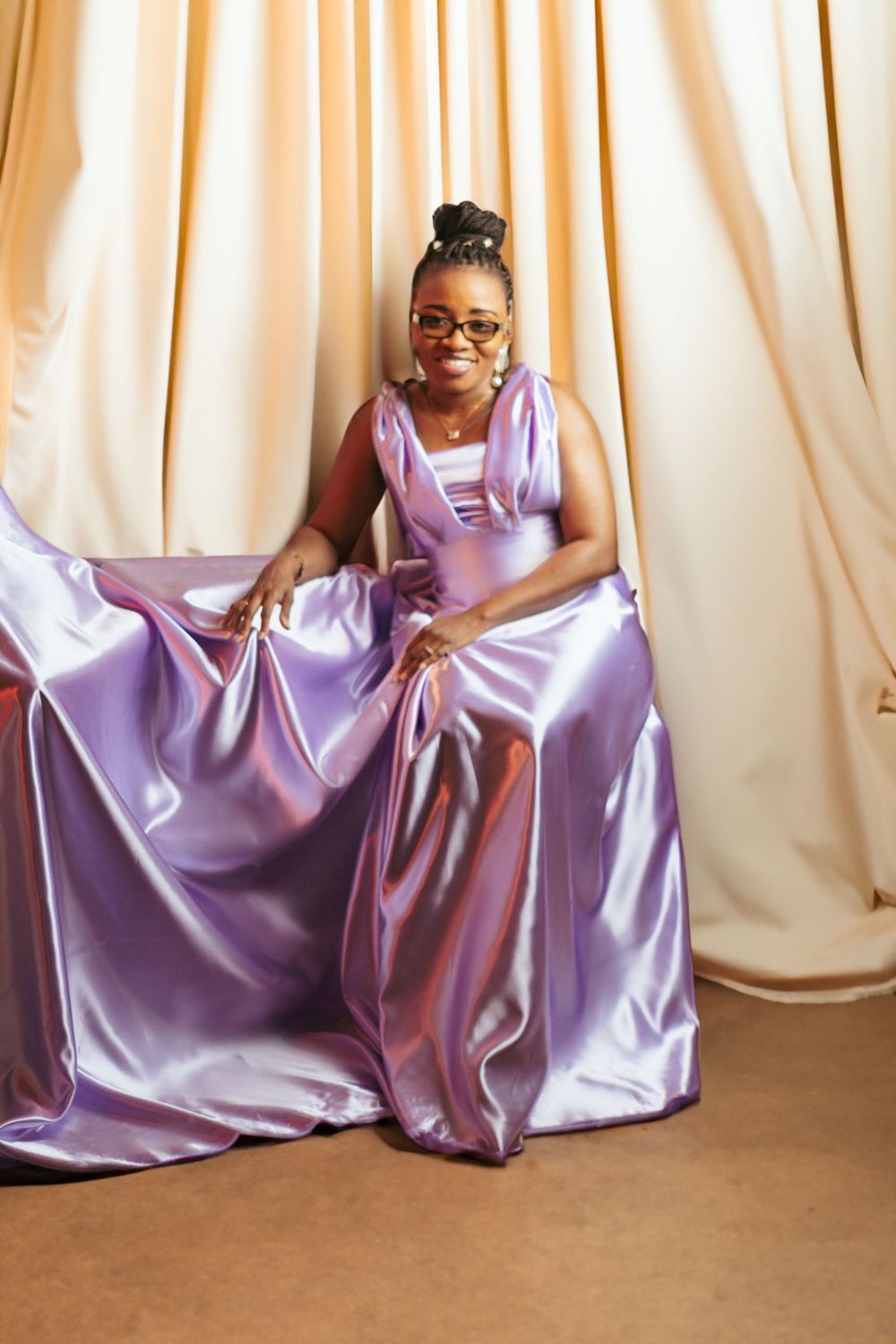 Person in a lavender satin dress poses elegantly against cream curtains.