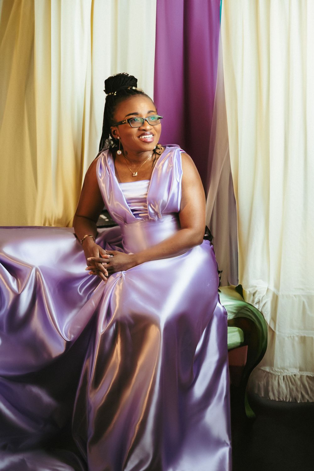 Model in glasses wearing a flowing lavender gown poses against draped curtains.