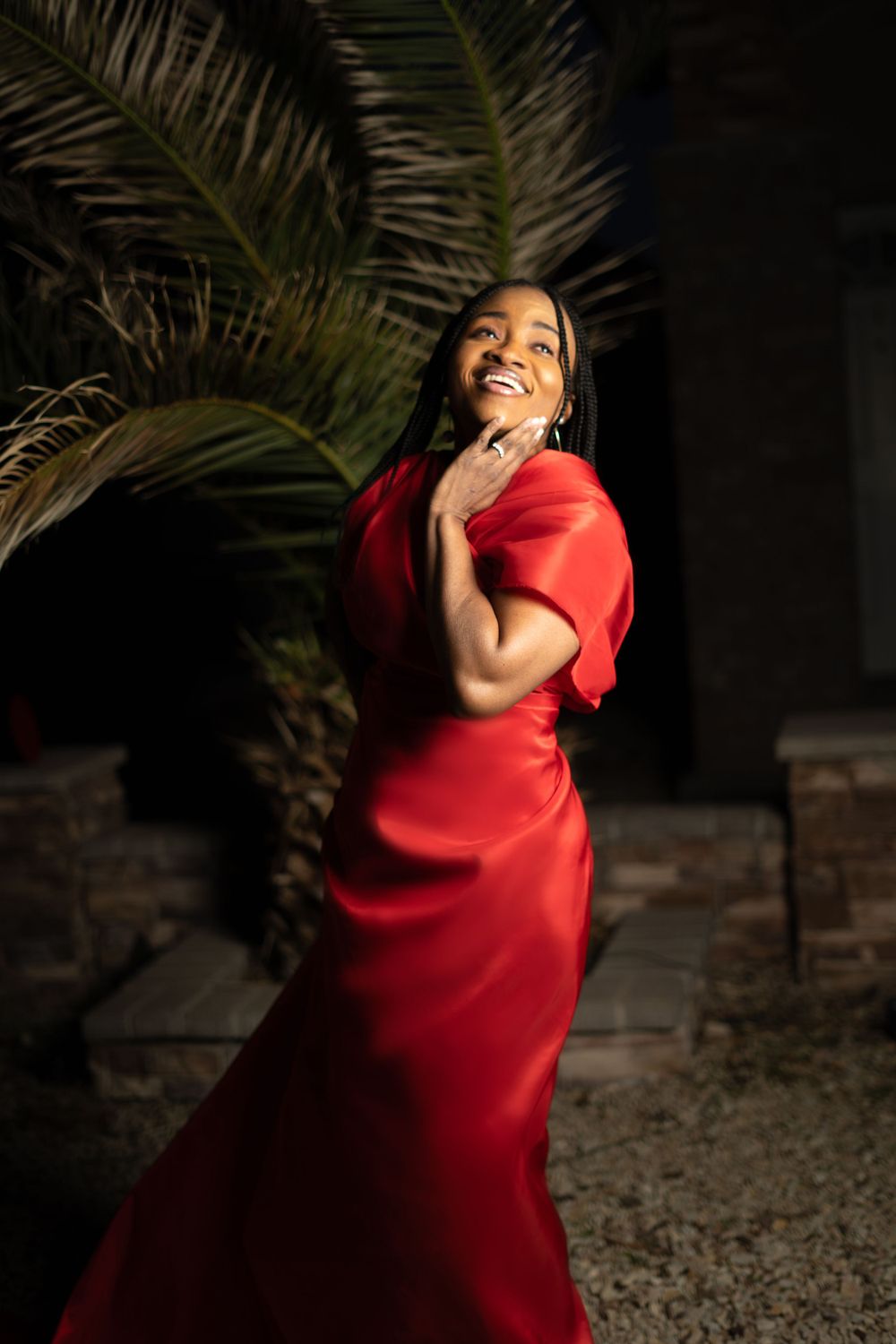 Person in striking red gown poses expressively near palm trees at night.