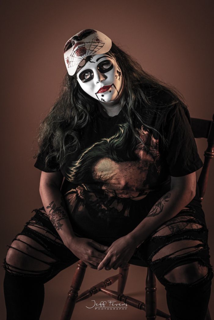 Woman sitting in chair in ripped jeans with a porcelain doll face mask on with another mask on top of her head.