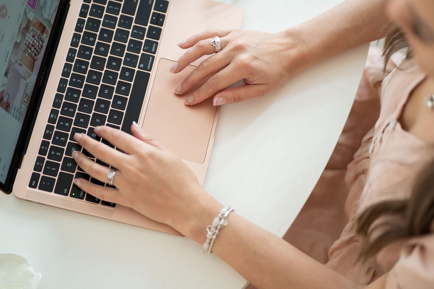 Hands typing on a rose gold laptop showing jewelry accessories and manicured nails.