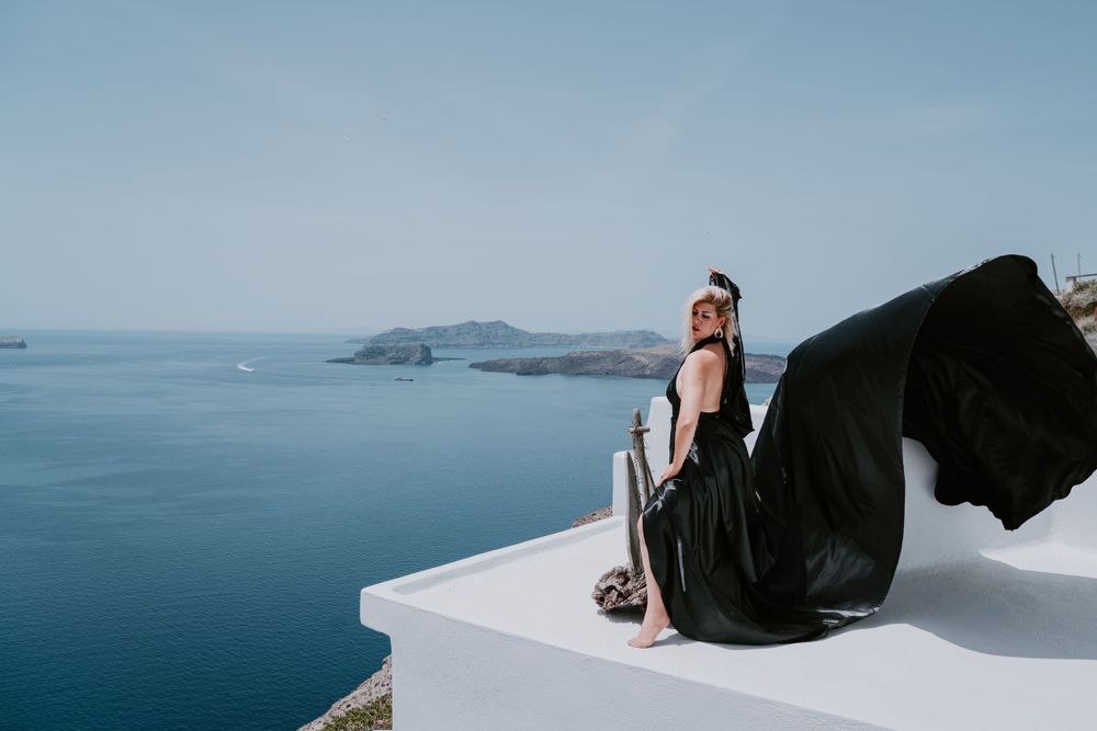 Flowing black dress fabric billows dramatically on a white terrace overlooking the Aegean Sea.