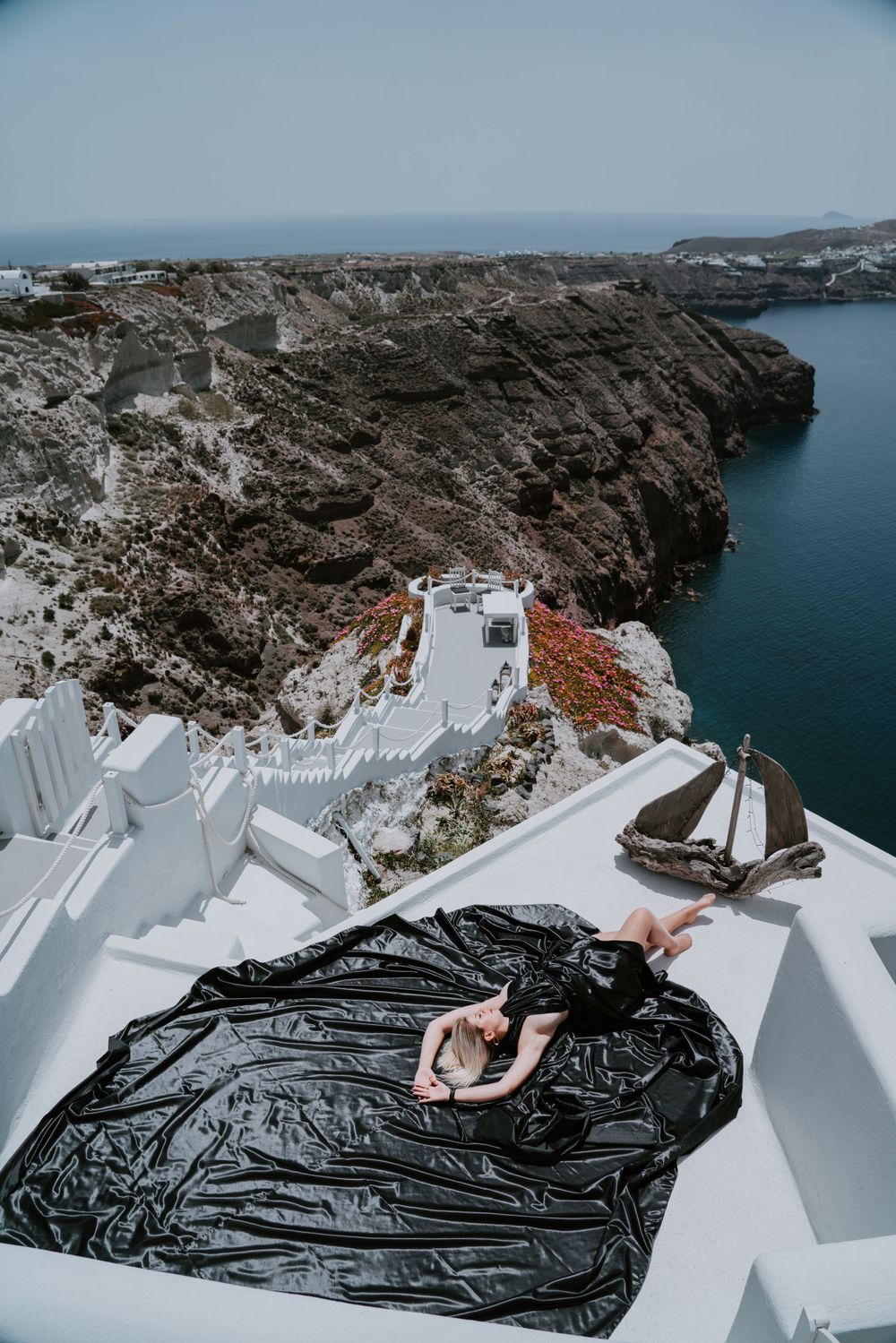 A dark fabric flows over white terraces overlooking dramatic cliffs and the Aegean Sea.