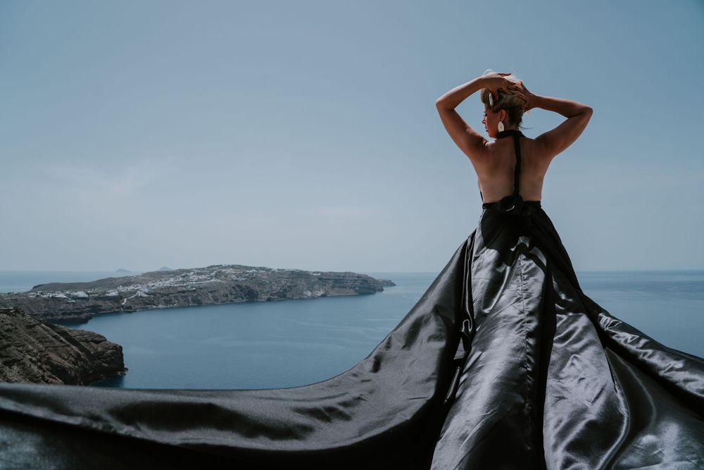 Dramatic fashion pose with black flowing dress against scenic Greek island vista.