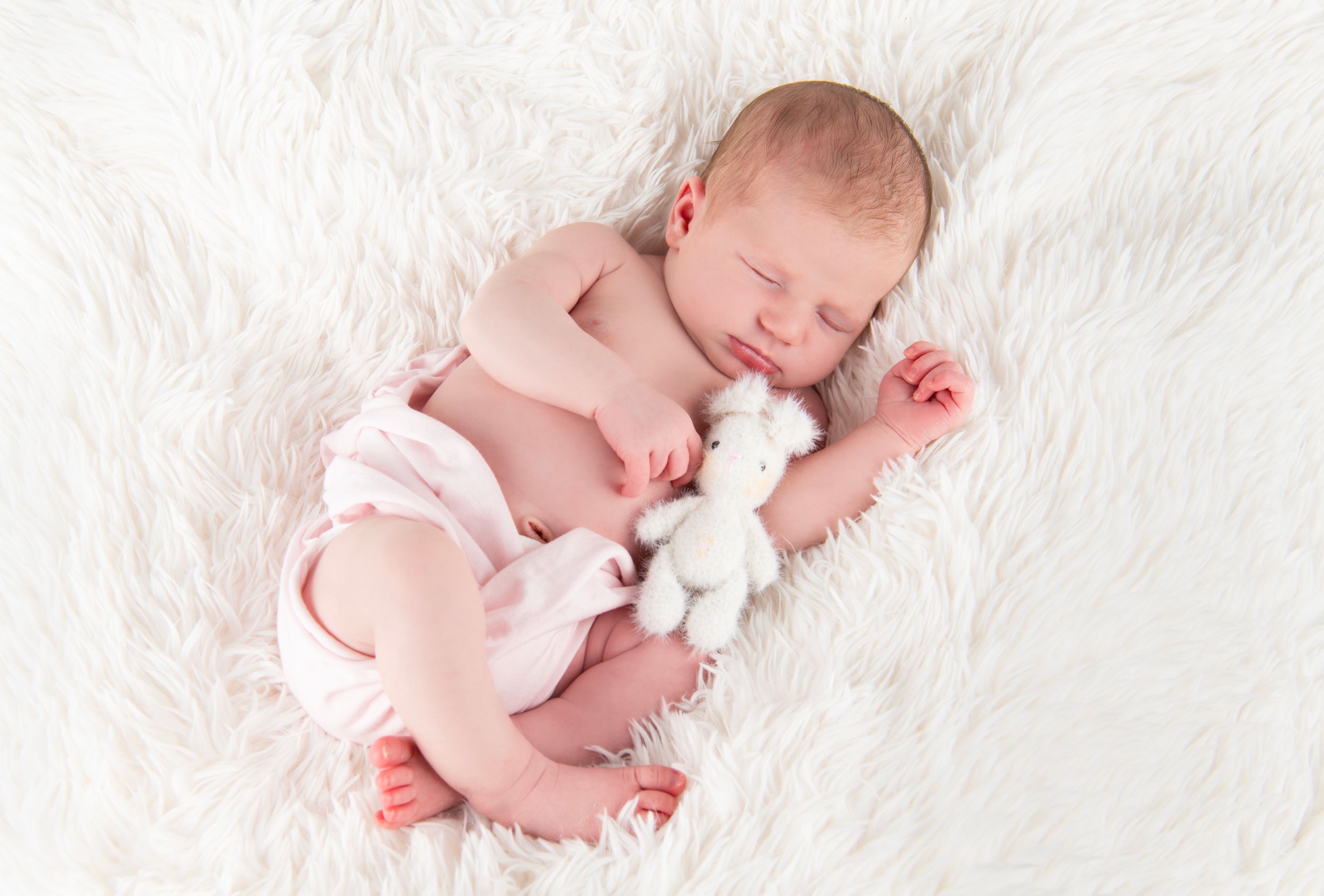 Sleeping newborn on white fur with small stuffed animal.