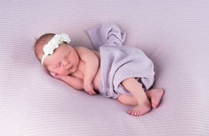 Sleeping newborn wearing white headband and purple wrap on textured blanket.