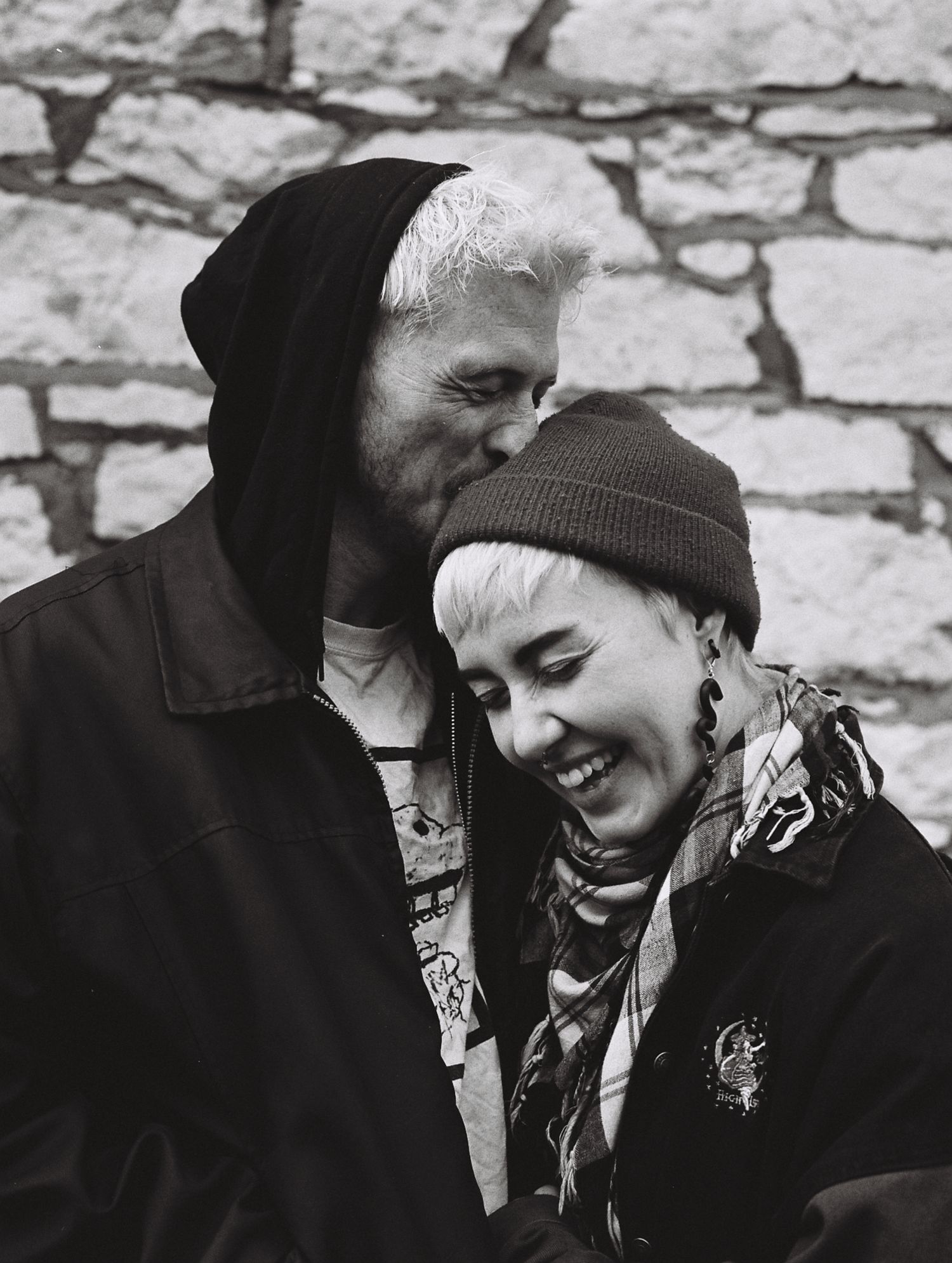 Black and white candid photo of a couple sharing a joyful moment against a stone wall.