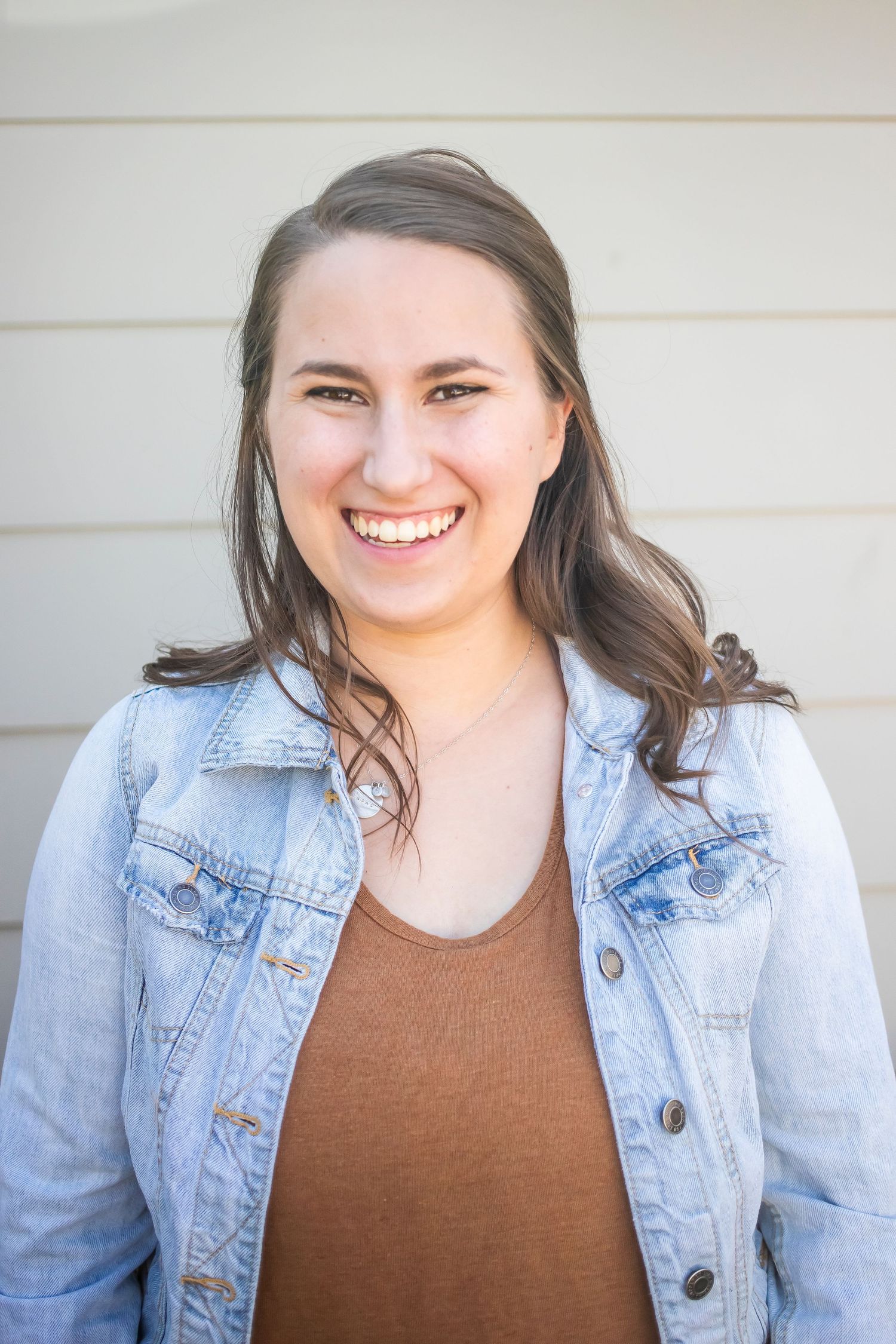 Portrait of someone wearing denim jacket and brown top against white siding.