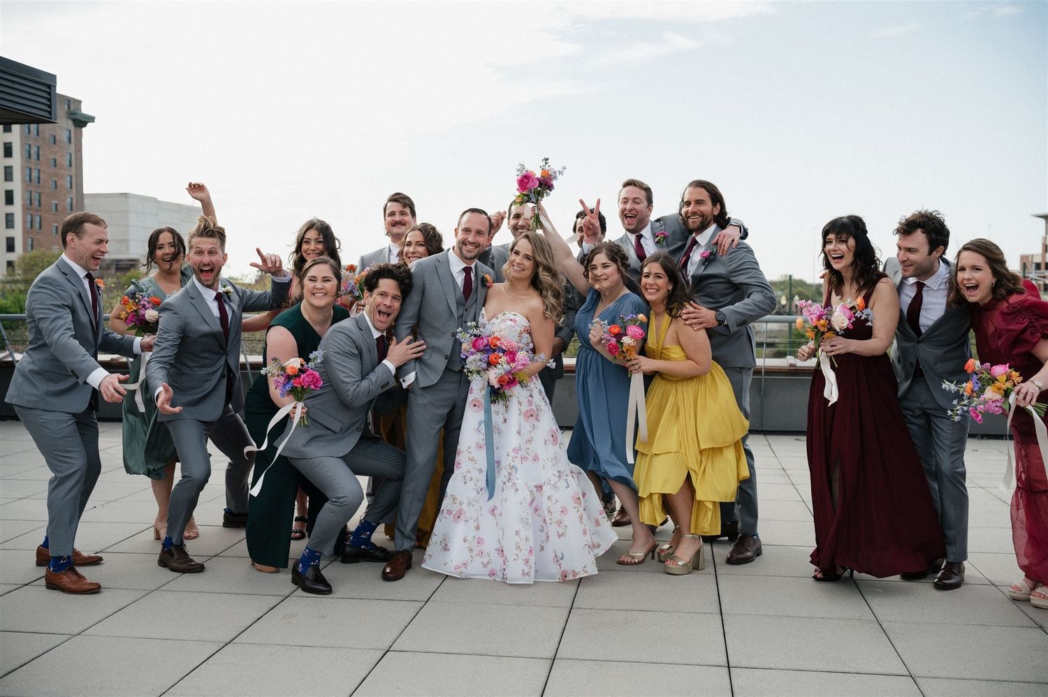 A wedding party in formal attire poses together on an outdoor rooftop terrace against a bright sky.