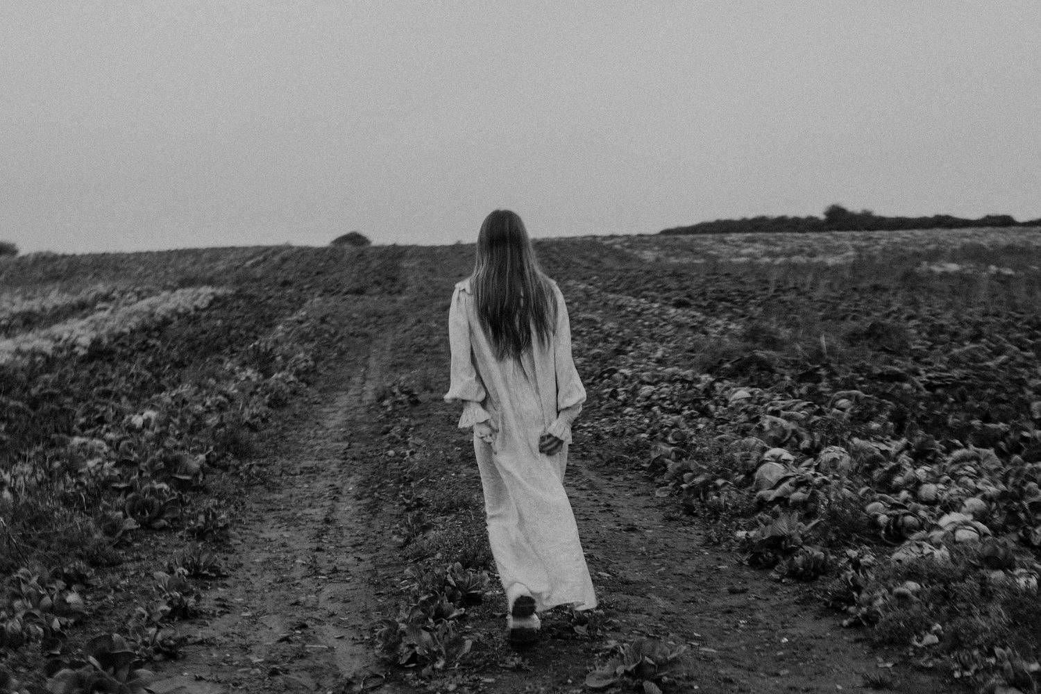 Person in flowing white dress walks alone down rural dirt path between crop fields.