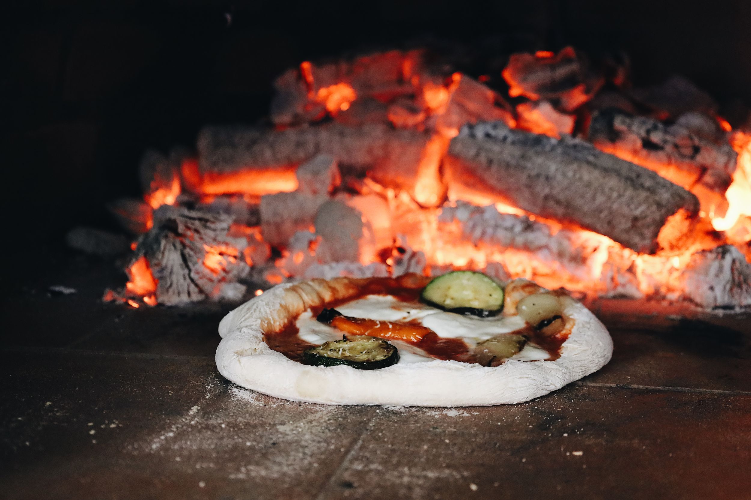 Wood-fired pizza cooking in a traditional brick oven with flames visible.