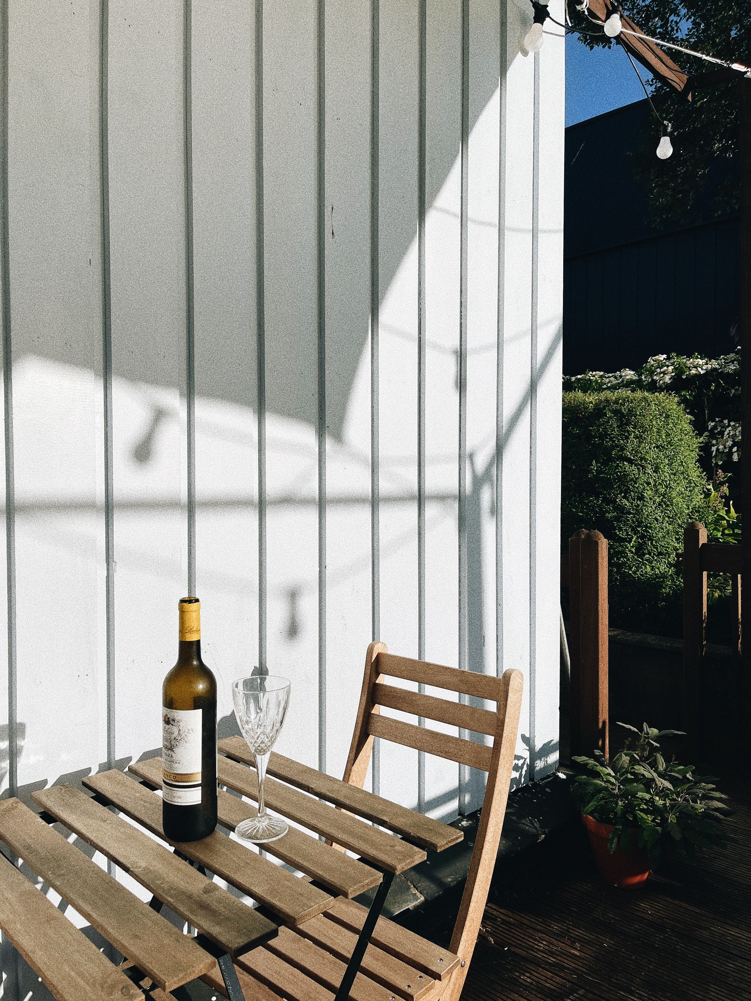 Outdoor dining setup with wooden bench and wine bottle casting shadows against white wall.