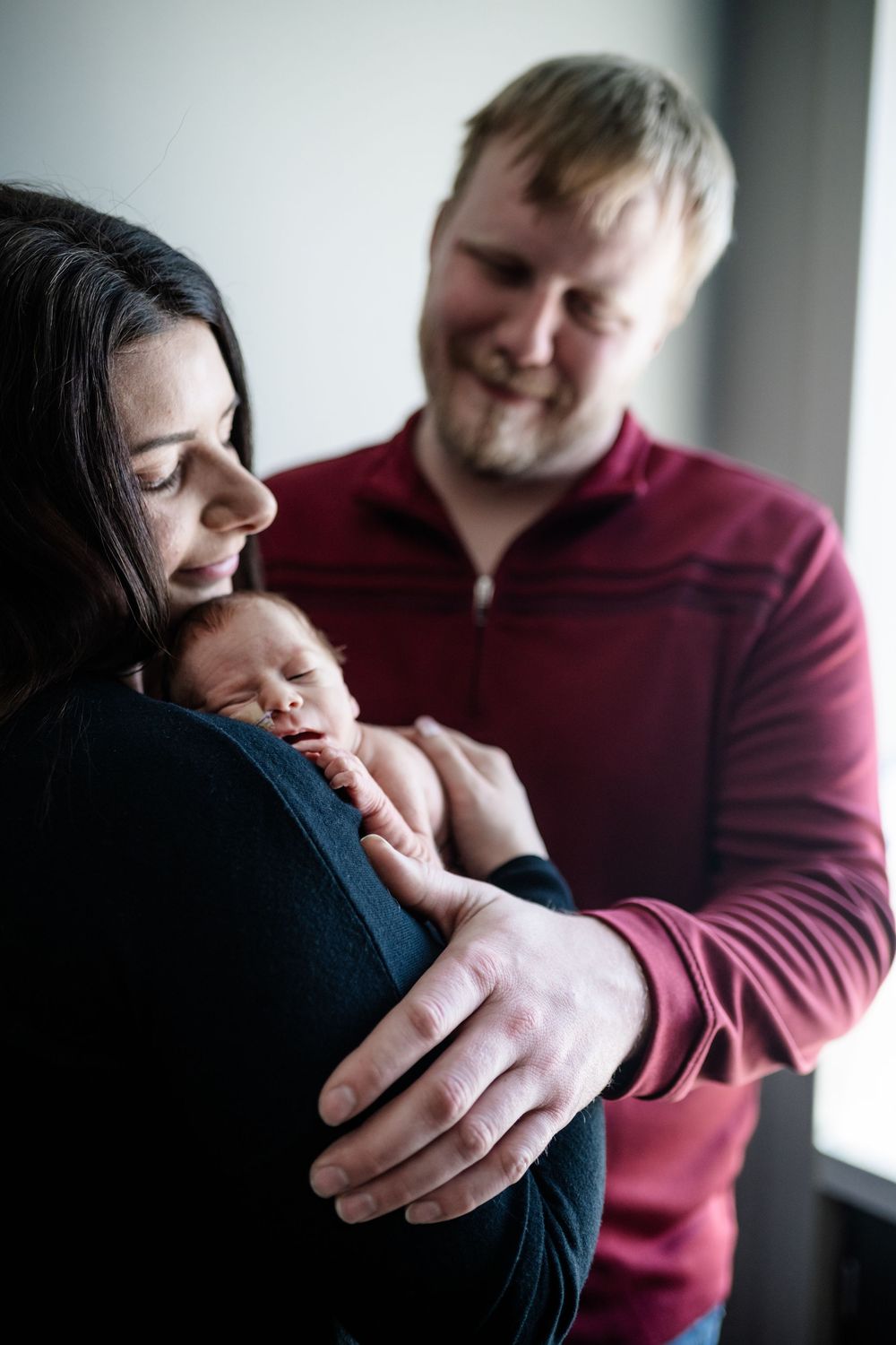 A couple shares a gentle moment with their newborn baby against a light background.