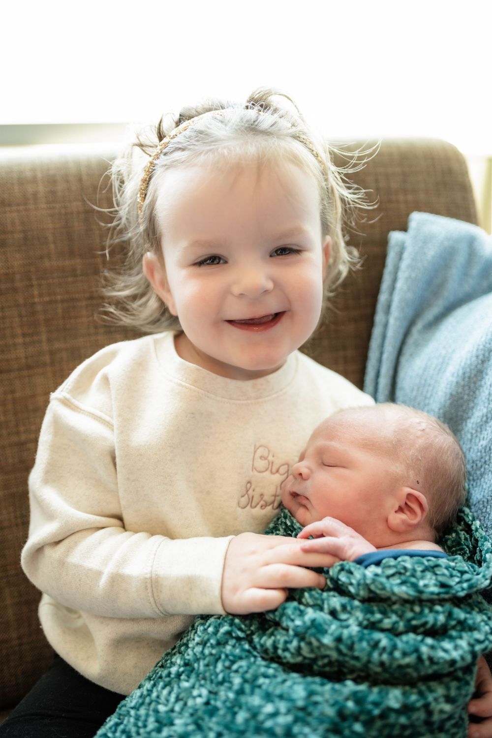 A toddler in a cream sweater smiles while looking at a swaddled newborn sibling.