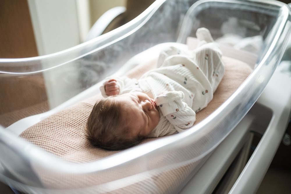 Newborn baby sleeping peacefully in a hospital bassinet, wrapped in a white onesie on a soft blanket.