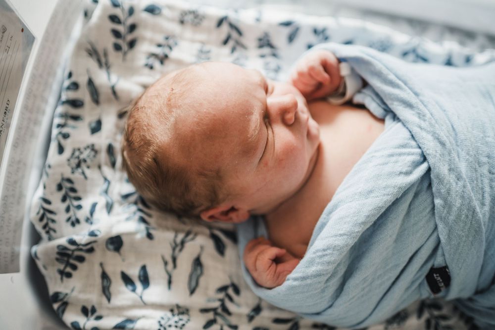 A sleeping infant wrapped in a grey swaddle rests peacefully on patterned bedding.