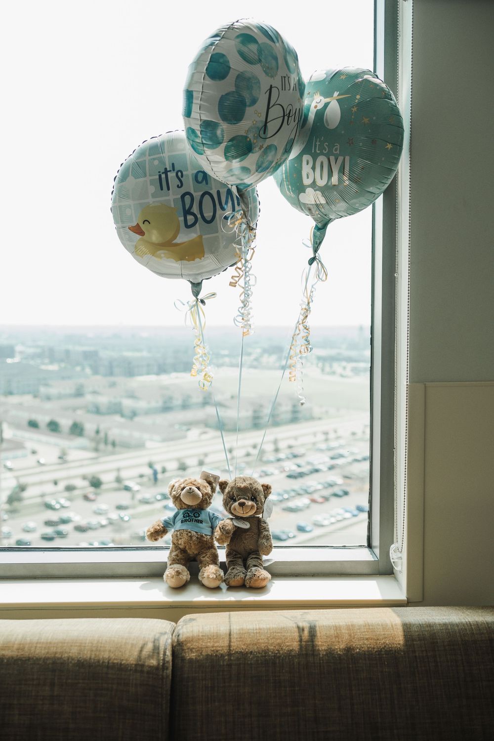 Two teddy bears sit on a windowsill with blue "boy" balloons floating above and a parking lot view in the background.