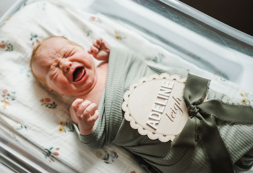 A crying baby lies on a white floral patterned sheet wearing a gray knit sweater.
