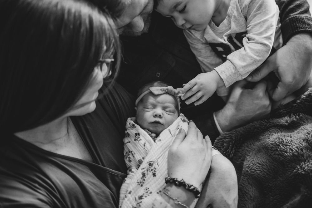 A black and white intimate photograph of a newborn baby swaddled in a patterned blanket being held by family members.