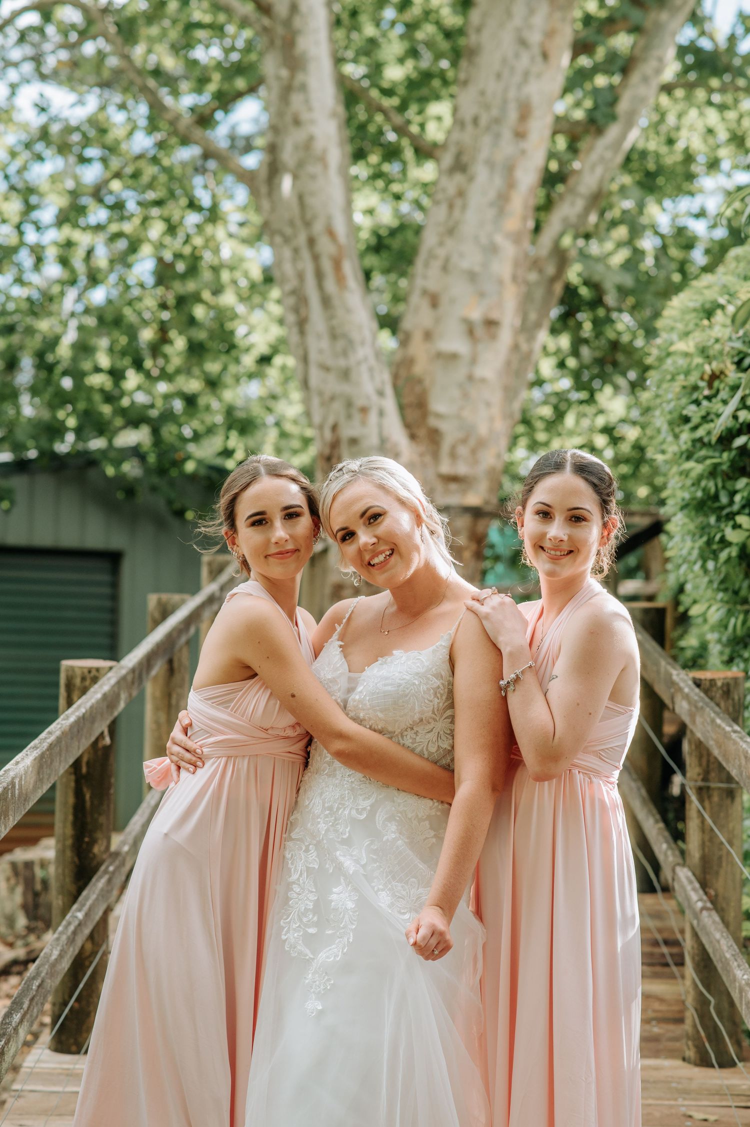 Bride and bridesmaids in pink dresses posing on a wooden bridge surrounded by trees.