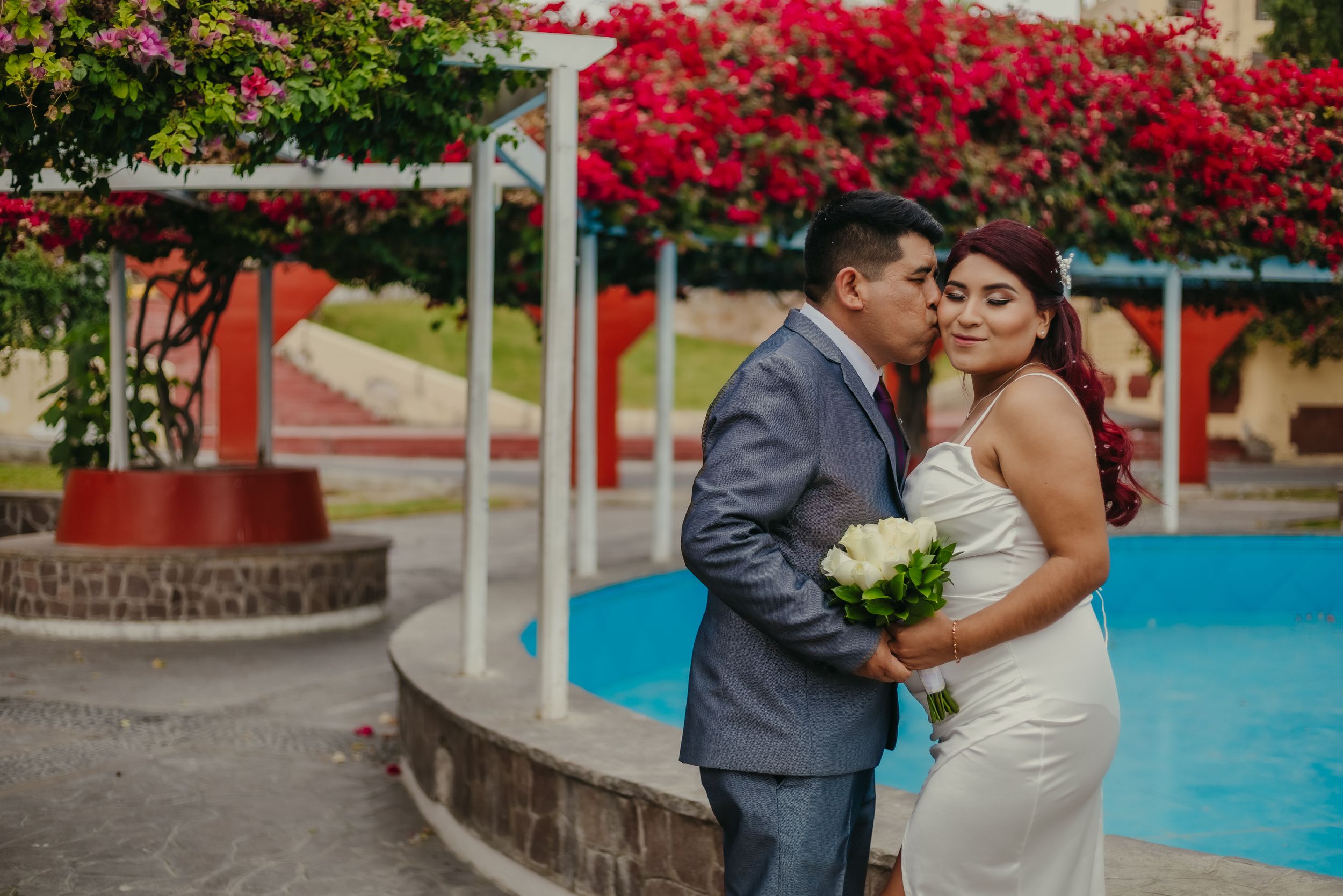 Couple shares intimate kiss in front of vibrant red bougainvillea flowers during wedding photoshoot.