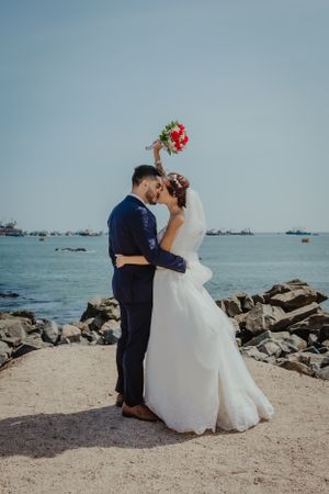 A romantic wedding couple embraces on a rocky beach with boats in the harbor background.
