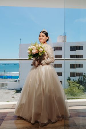 Bride in lace wedding gown holding pink bouquet on balcony overlooking ocean.