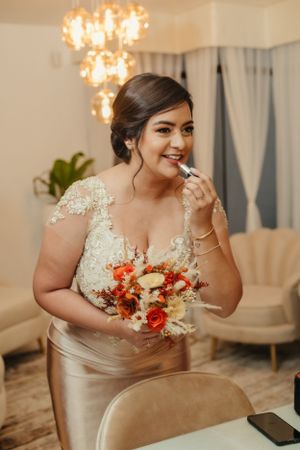 Bride holds autumn-colored bouquet while applying makeup in elegant room setting.