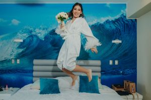 Bride in white robe jumps joyfully on bed against blue ocean wave wall mural holding bouquet.