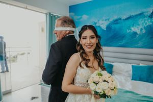 Bride holding white bouquet poses in wedding dress against ocean-themed backdrop.