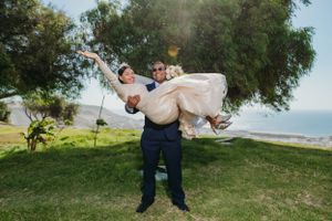 Groom carrying bride playfully on grassy oceanside hill.