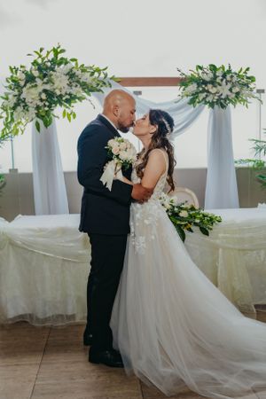 Couple shares romantic kiss under floral arch with white draping during wedding ceremony.